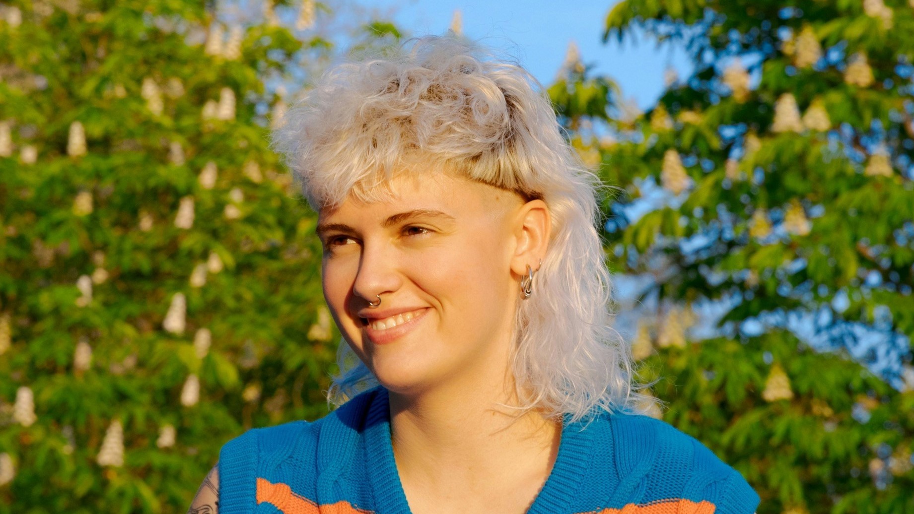 Una giovane donna con un mullet riccio biondo platino, seduta all'aperto in una giornata di sole.