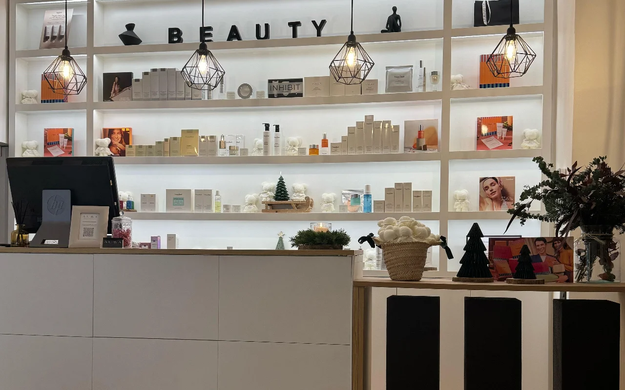 Una luminosa area reception moderna con un grande scaffale bianco retroilluminato pieno di prodotti per la cura della pelle. Dal soffitto pendono lampade geometriche a gabbia nera, e il bancone è un desk bianco e minimalista.