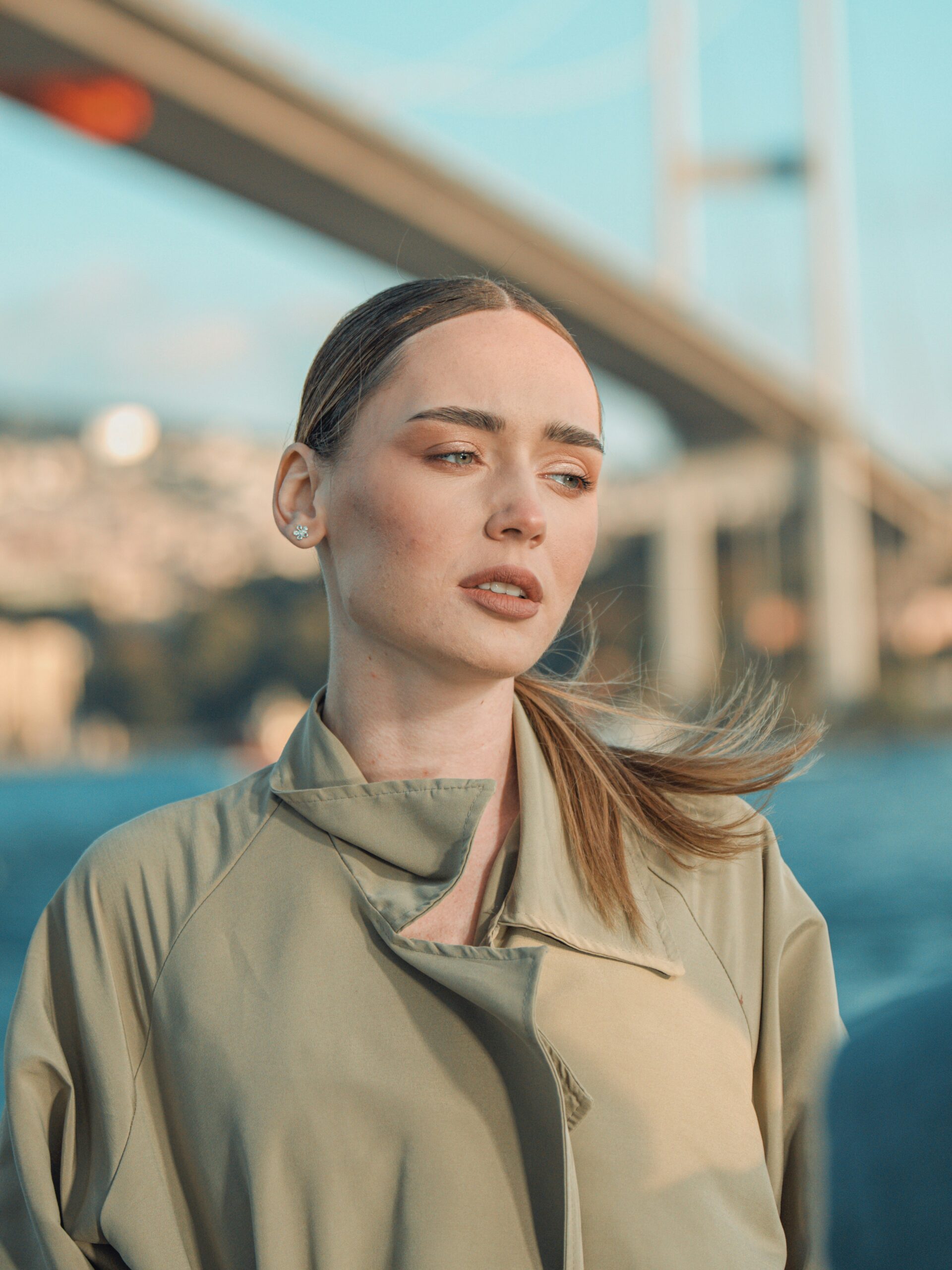 Una donna con microblading sulle sopracciglia e capelli chiari raccolti, trench coat, in piedi davanti a un ponte a Istanbul