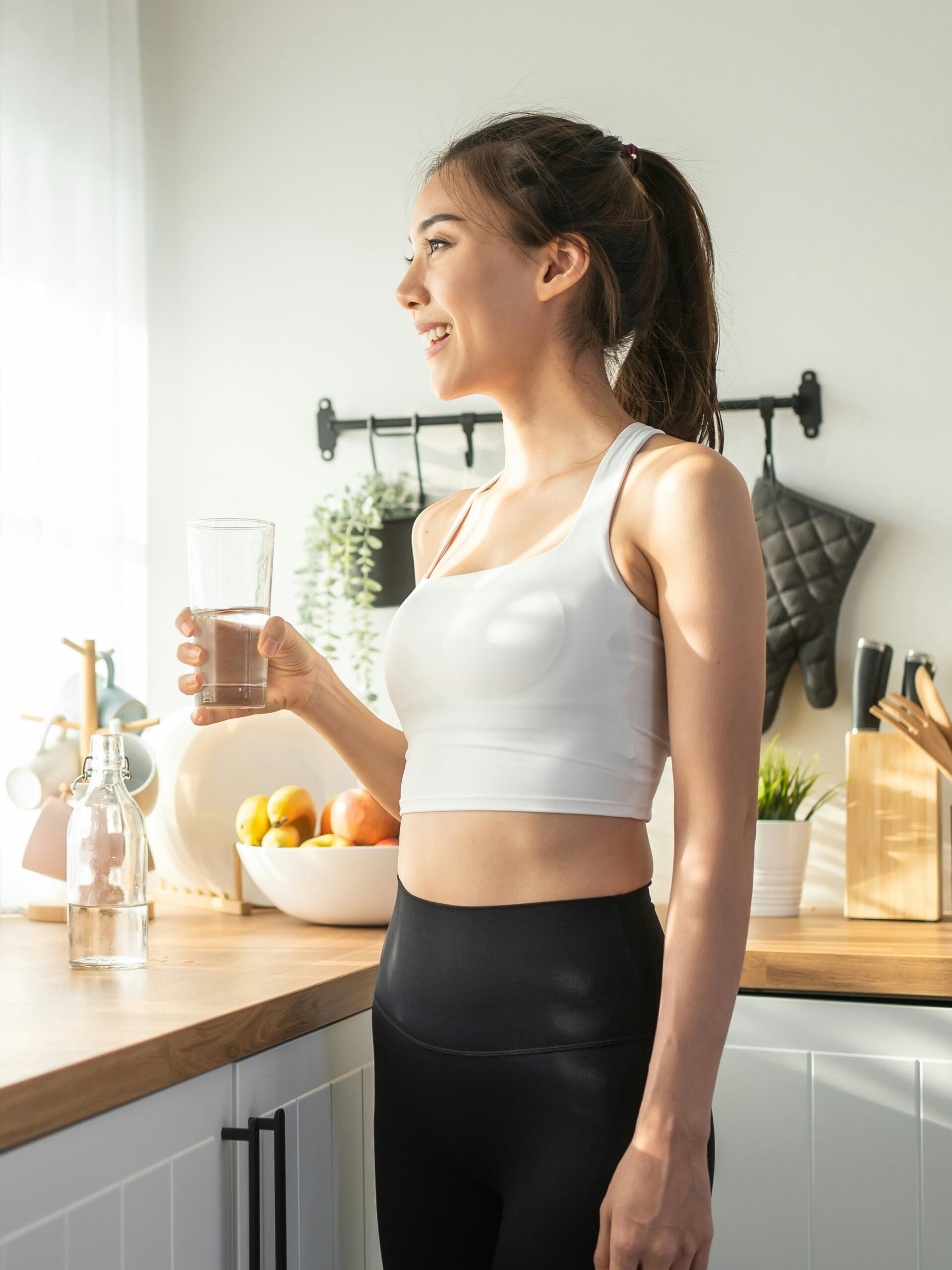 Vista laterale di una donna che beve un bicchiere d’acqua in cucina, davanti a una finestra soleggiata