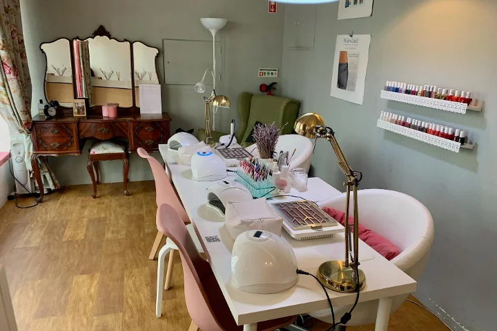 Vista interna di un accogliente salone di bellezza specializzato in manicure. In primo piano, un lungo tavolo bianco è allestito con lampade per unghie, luci da tavolo e vari strumenti da onicotecnica, con sedie rosa e bianche ai lati. Sullo sfondo, una toeletta in legno vintage con specchio a tre ante, una poltrona verde e scaffali bianchi a parete con file di smalti colorati.