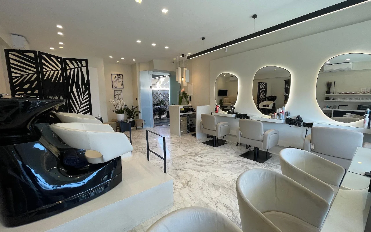 Un salone di parrucchiere elegante con pavimenti in marmo bianco, poltrone per lo styling in pelle crema sotto specchi ovali con cornice a LED, una postazione lavaggio nera lucida e un divisorio geometrico con motivo a foglia di palma.