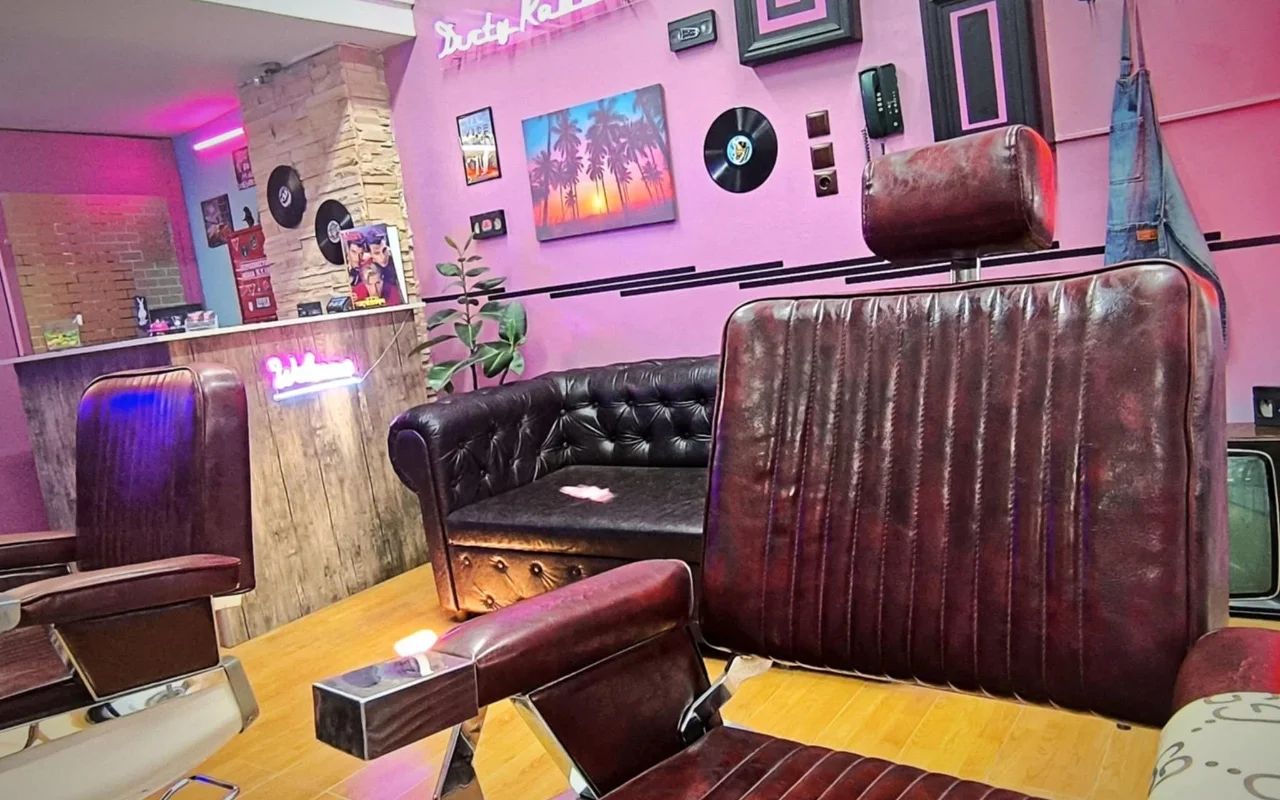 Una barberia in stile retrò con pareti rosa e viola, insegne al neon, dischi in vinile usati come decorazione, un dipinto con palma e tramonto, poltrone da barbiere in pelle scura e un divano Chesterfield su un pavimento in legno caldo.