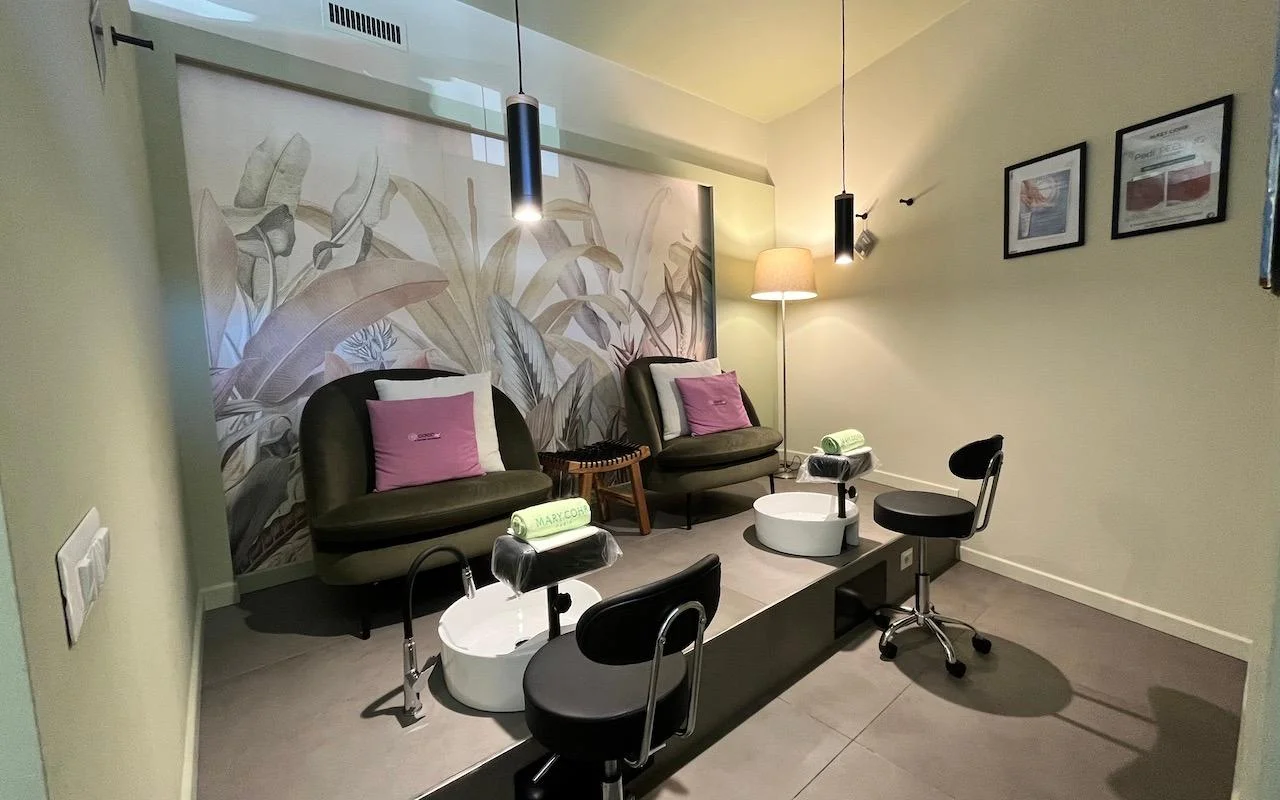 Una postazione pedicure raffinata con due poltrone in velluto verde intenso e cuscini rosa. Vasche per i piedi bianche sono incassate in una piattaforma grigia rialzata, su sfondo di un murale con grandi foglie tropicali.