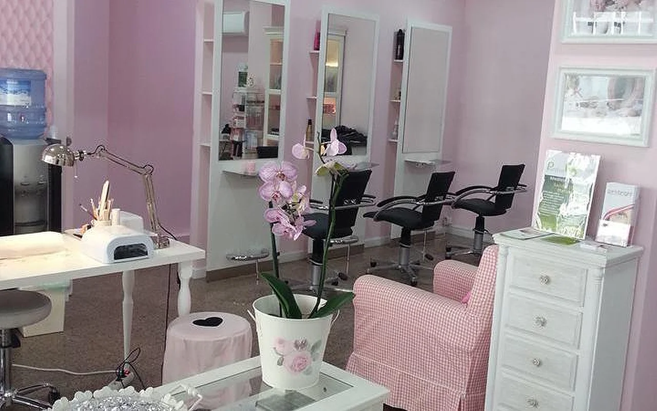 L'interno di un salone luminoso a tema rosa dedicato a capelli e unghie. Sulla sinistra, una postazione manicure bianca è attrezzata con lampada da tavolo e dispositivo polimerizzante. Al centro, un'orchidea in un vaso bianco poggia su un tavolino da caffè accanto a una poltroncina a quadretti rosa e bianca. Sullo sfondo, diverse postazioni da parrucchiere con specchi alti e poltrone nere contro pareti rosa chiaro.