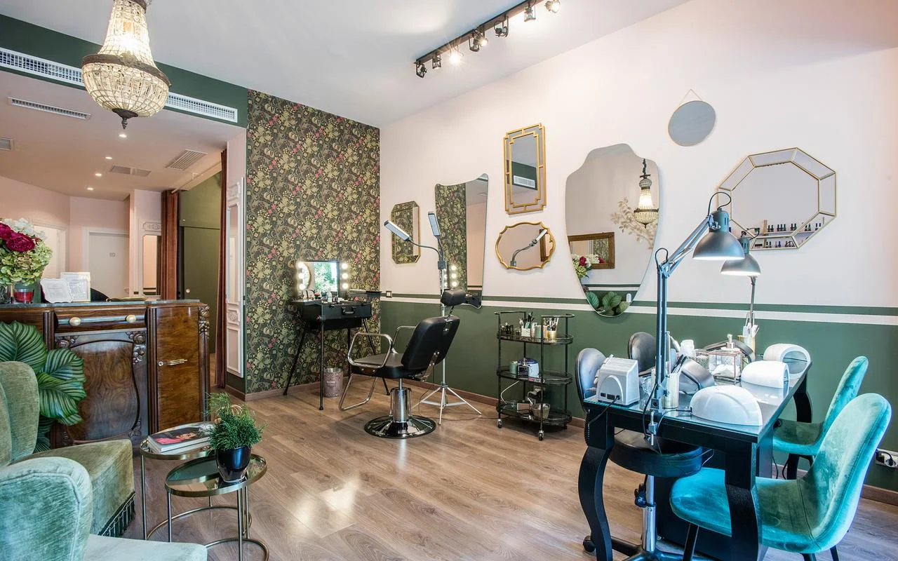 Un salone dallo stile vintage-chic con pareti bianche e verde bosco. Presenta carta da parati floreale, un assortimento di specchi con cornici dorate irregolari, una postazione make-up professionale e sedute in velluto verde.