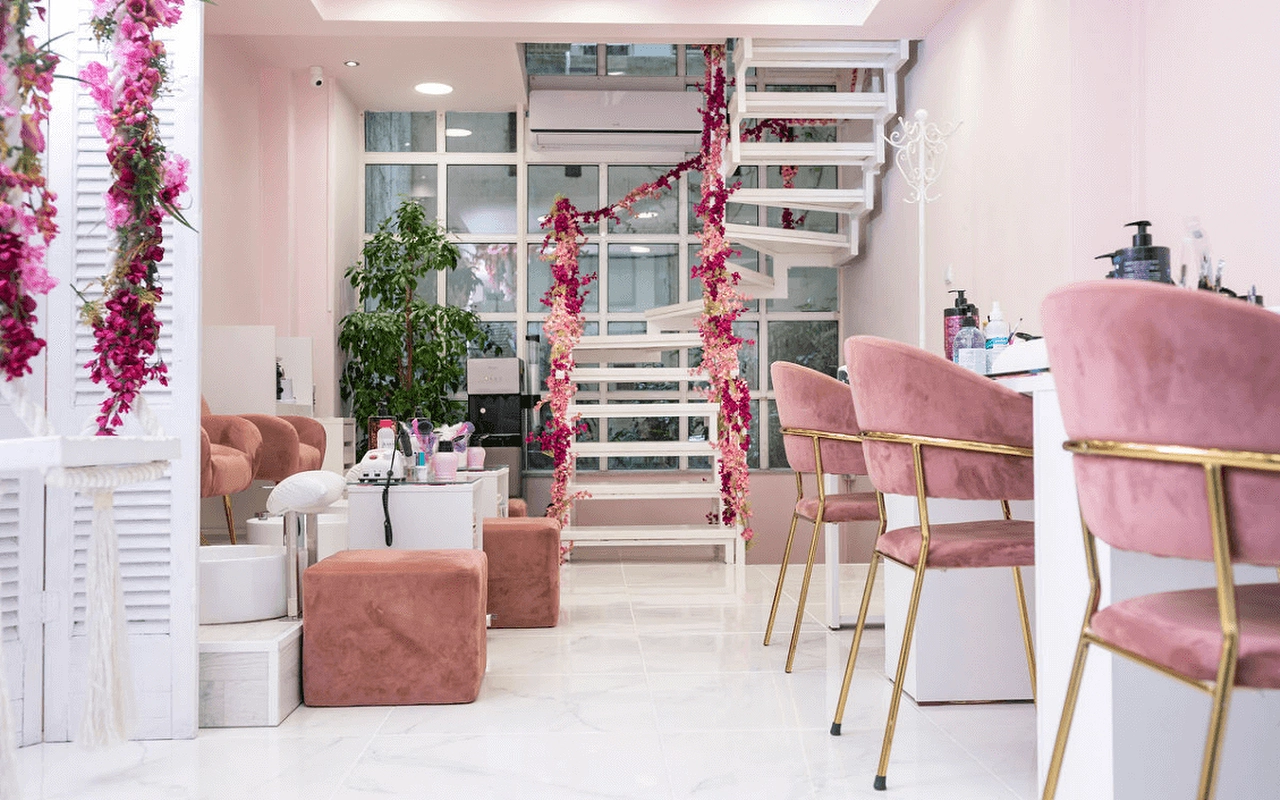 Un salone unghie dall'atmosfera femminile con poltrone in velluto rosa cipria, una scala a chiocciola bianca drappeggiata di fiori finti rosa, divisori a persiana, postazioni manicure con gambe dorate su un pavimento effetto marmo.