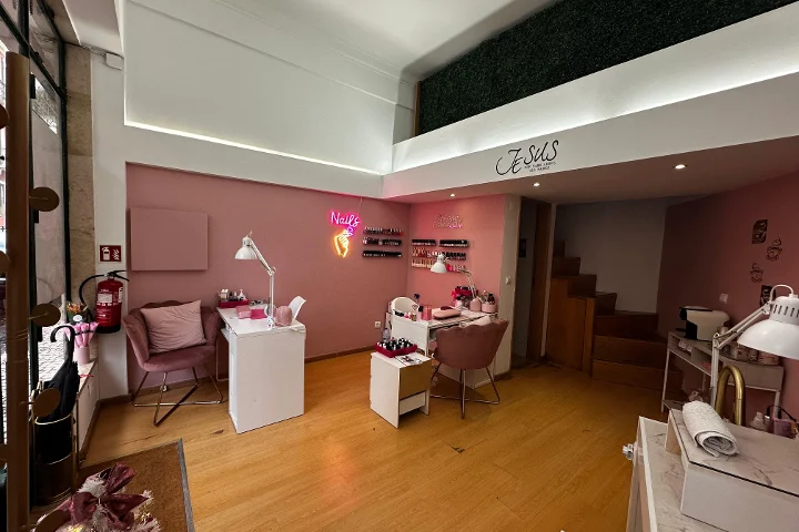 Un elegante studio per unghie con pareti rosa chiaro e pavimenti in legno caldo. La stanza contiene diversi tavoli da manicure bianchi abbinati a poltroncine in velluto rosa antico. Una luminosa insegna al neon rosa a forma di mano con uno smalto e la scritta 'Nails' brilla sulla parete, e file di smalti sono esposti su piccole mensole. Un soppalco con una parete di siepe finta verde è visibile in alto.