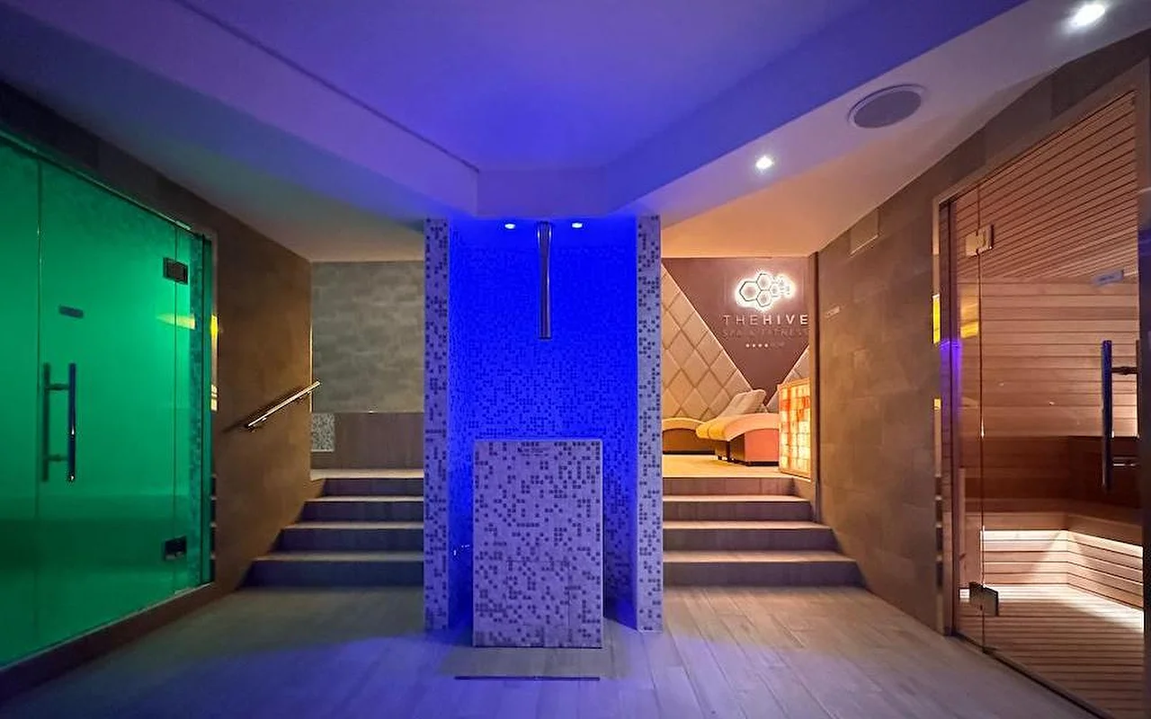 L'area termale di una spa di lusso con un'illuminazione scenografica blu e verde. Lo spazio comprende una sauna finlandese in legno, l'ingresso al bagno turco e una zona centrale con mosaici e gradini che conducono alle aree relax.