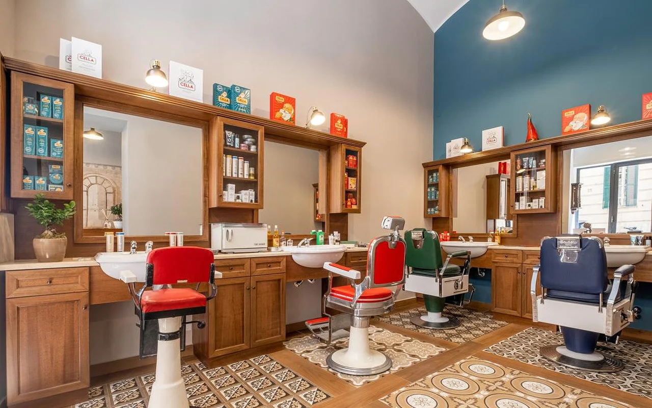 Una barberia tradizionale di alta gamma con mobili in legno pregiato, piastrelle a pavimento dallo stile vintage e poltrone da barbiere professionali in rosso, verde e blu navy. Prodotti da barba tradizionali sono esposti sugli scaffali.