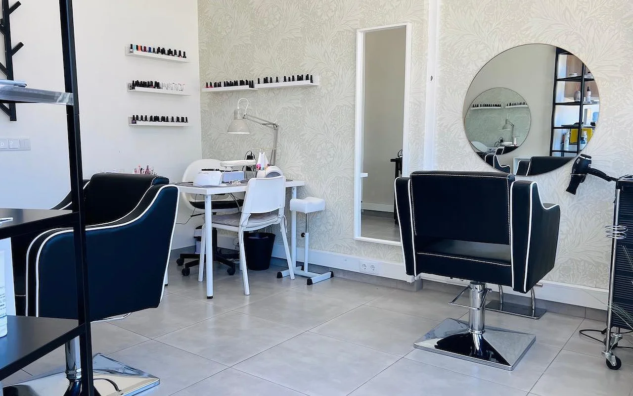 Un compatto salone di capelli e unghie con poltrone da styling nere profilate di bianco, uno specchio rotondo e uno specchio alto su una carta da parati grigia a fiori, e mensole a parete allineate di smalti.