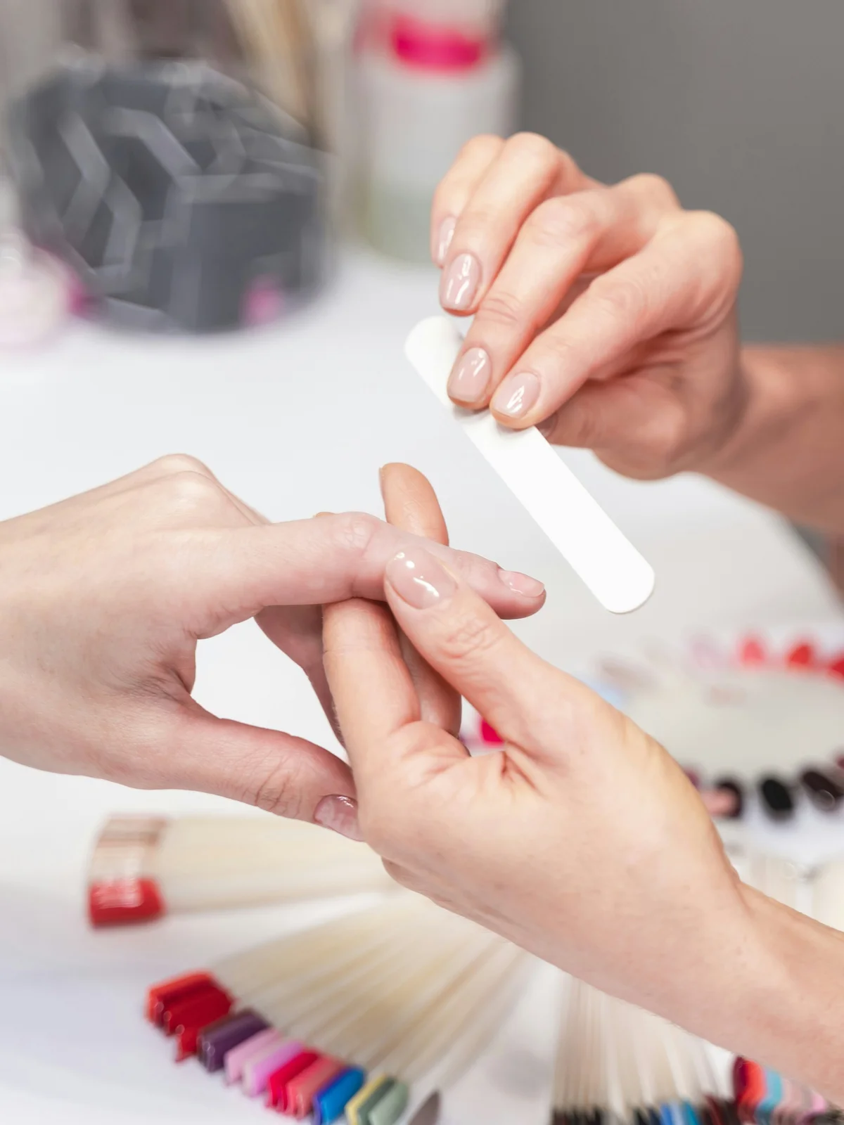 Primo piano di una manicurista che lima delicatamente le unghie di una cliente in salone, con prodotti e strumenti per manicure sullo sfondo